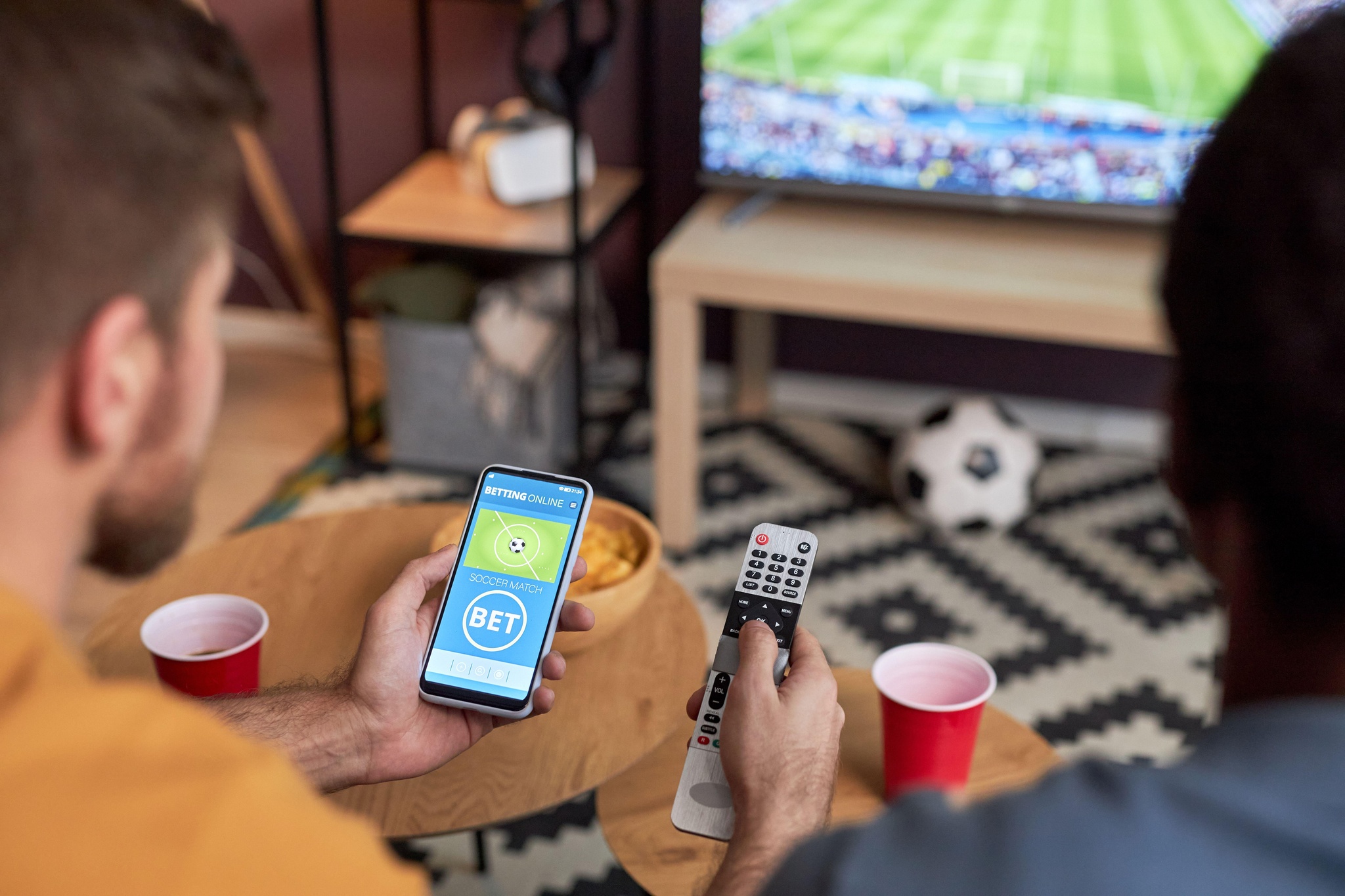 Close up of two men watching football match on TV and holding smartphone with online sports bets